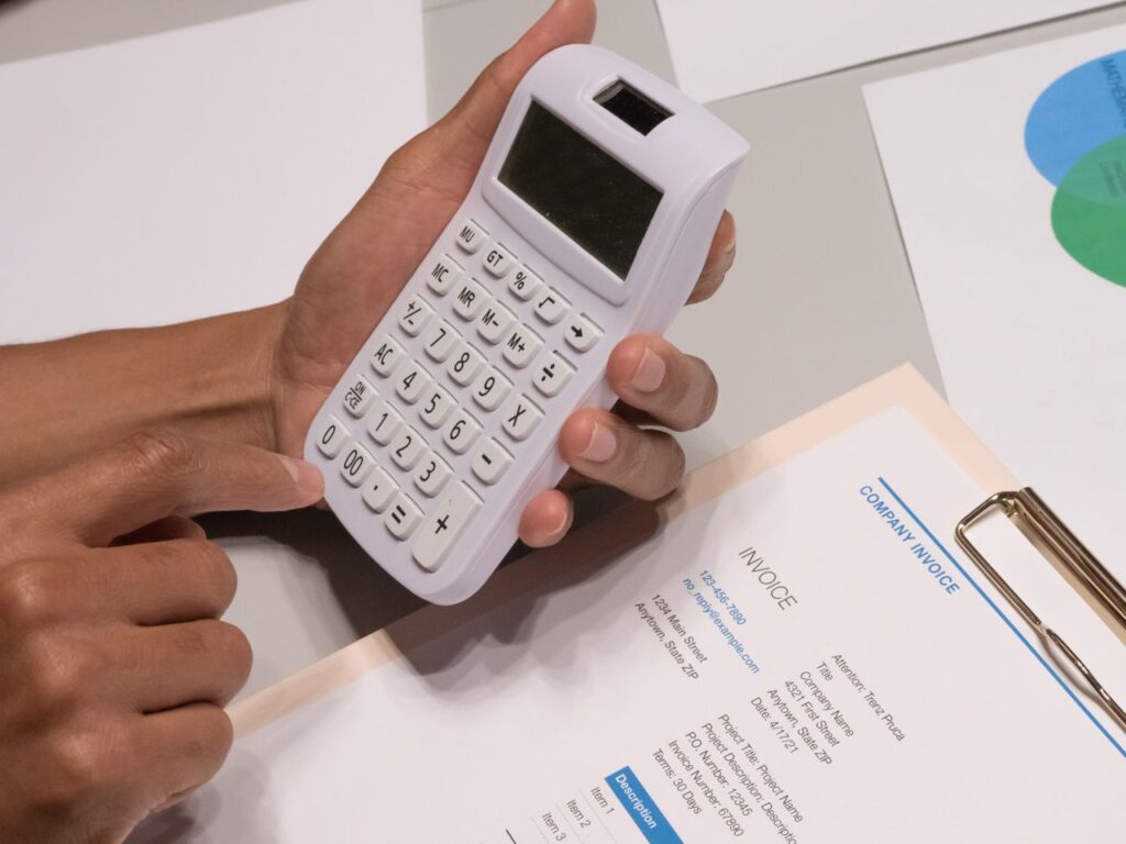 pexels-photo-7688524-7688524 Close-up of hands using a calculator next to a company invoice, depicting a financial calculation concept.